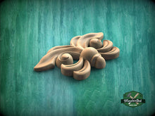 Load image into Gallery viewer, A wooden carving of a stylized flower or leaf design is displayed against a green background.