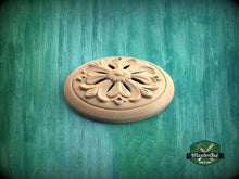 Load image into Gallery viewer, A wooden ornamental piece with a floral design sits on a green textured background.
