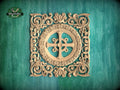 a square, ornately carved wooden cross with intricate floral designs surrounding it, set against a teal background.
