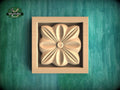 A wooden relief in the shape of a flower is displayed in a square frame against a green background.