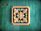 a square wooden carving with an intricate floral design, set against a green background.